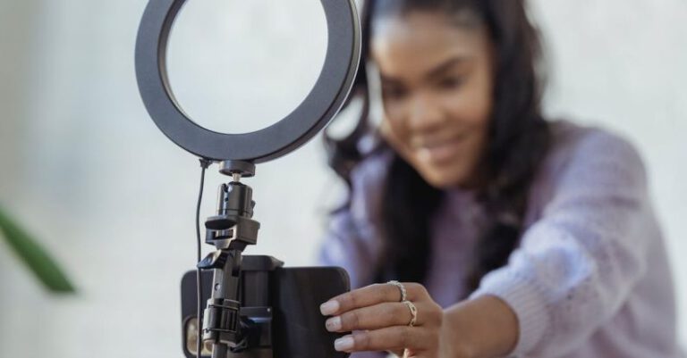 Tutorials - Cheerful young African American female blogger in stylish sweater smiling while setting up camera of smartphone attached to tripod with ring light before recording vlog