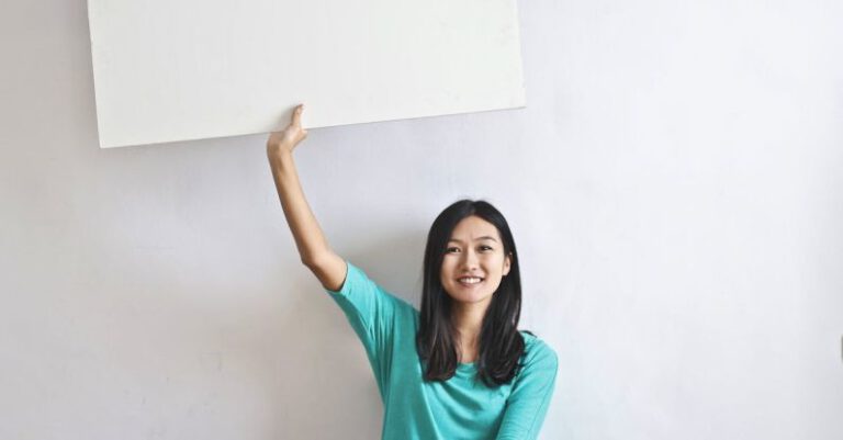 Promote - Cheerful Asian woman sitting cross legged on floor against white wall in empty apartment and showing white blank banner